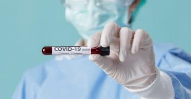 Profissional de saúde usando luvas e máscara, enquanto segura um pequeno frasco com os escritos "COVID-19"