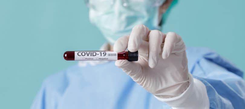 Profissional de saúde usando luvas e máscara, enquanto segura um pequeno frasco com os escritos "COVID-19"
