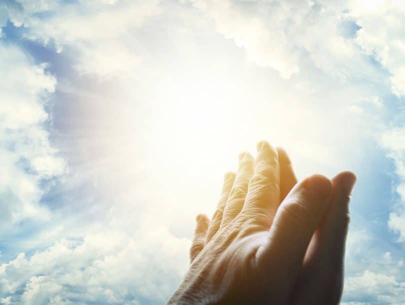 Mãos unidas em sinal de oração, apontando para um céu ensolardo.