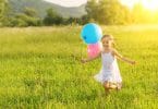 Menina pequena correndo feliz por um campo, enquanto segura balões de aniversário.