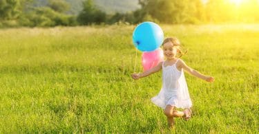 Menina pequena correndo feliz por um campo, enquanto segura balões de aniversário.