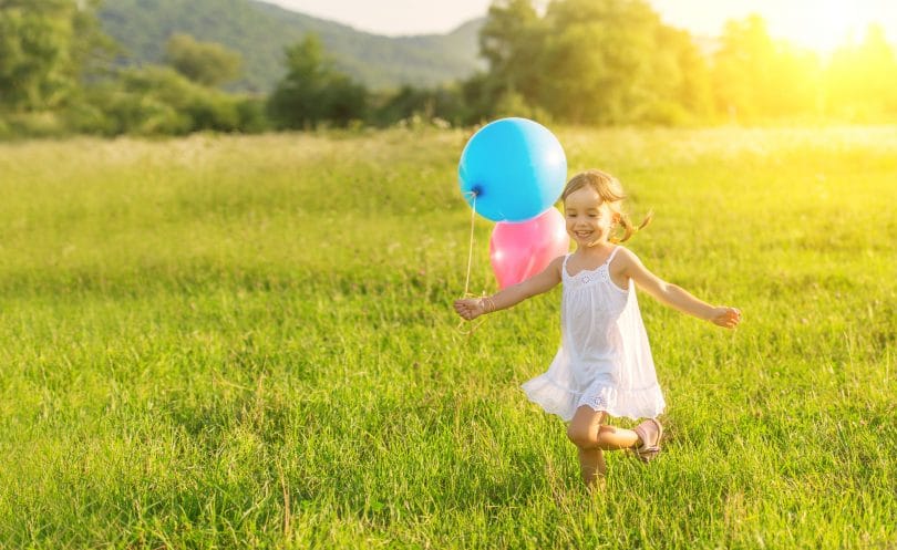 Menina pequena correndo feliz por um campo, enquanto segura balões de aniversário.