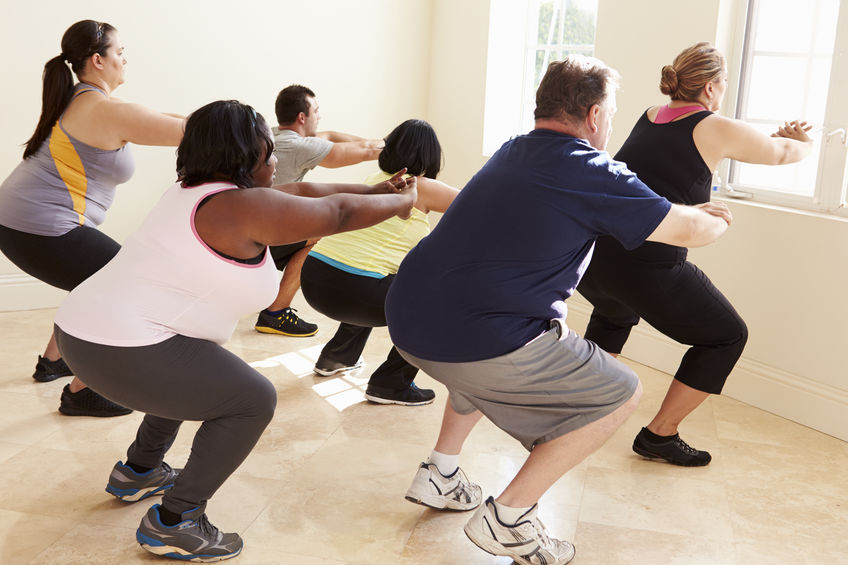 Pequeno grupo de pessoas obesas observando uma treinadora ensinando um exercício enquanto o grupo reproduz.