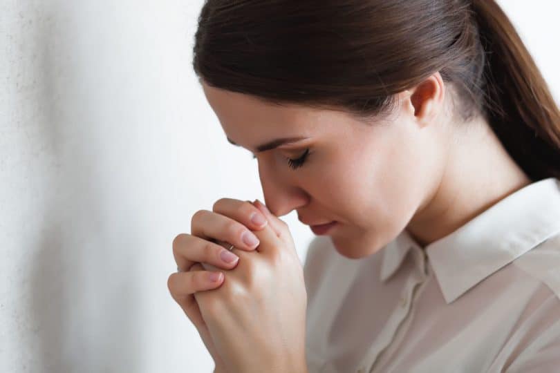 Mulher de olhos fechados e cabeça baixa, com as mãos entrelaçadas em sinal de oração.