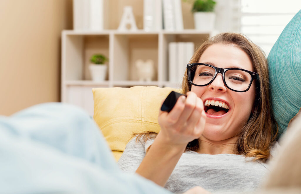 Mulher rindo deitada no sofá olhando para a TV