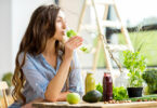 Mulher de cabelos longos e ondulados sentada em uma mesa de madeira, bebendo água aromatizada com limão e ervas. Sobre a mesa, estão vários tipos de hortaliças, sucos naturais e frutas.