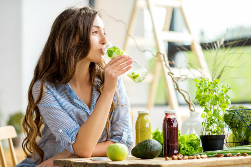 Mulher de cabelos longos e ondulados sentada em uma mesa de madeira, bebendo água aromatizada com limão e ervas. Sobre a mesa, estão vários tipos de hortaliças, sucos naturais e frutas.