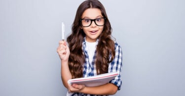 Garotinha usando óculos com caneta e caderno na mão com expressão de surpresa