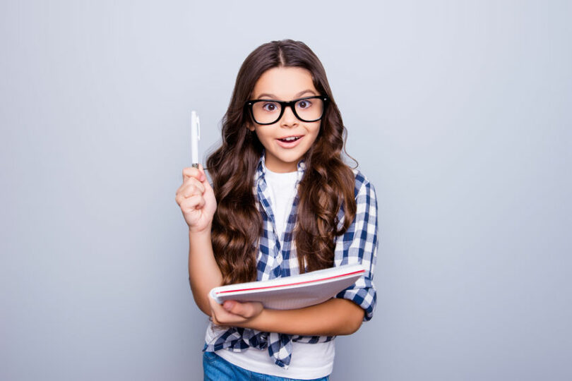 Garotinha usando óculos com caneta e caderno na mão com expressão de surpresa