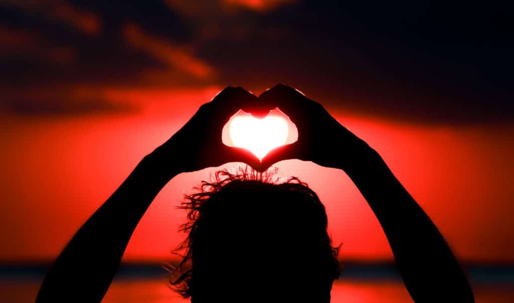 Imagem de um homem com os braços erguidos. As suas mãos estão juntadas e formam a imagem de um coração. Ao fundo temos a imagem do pôr do sol.
