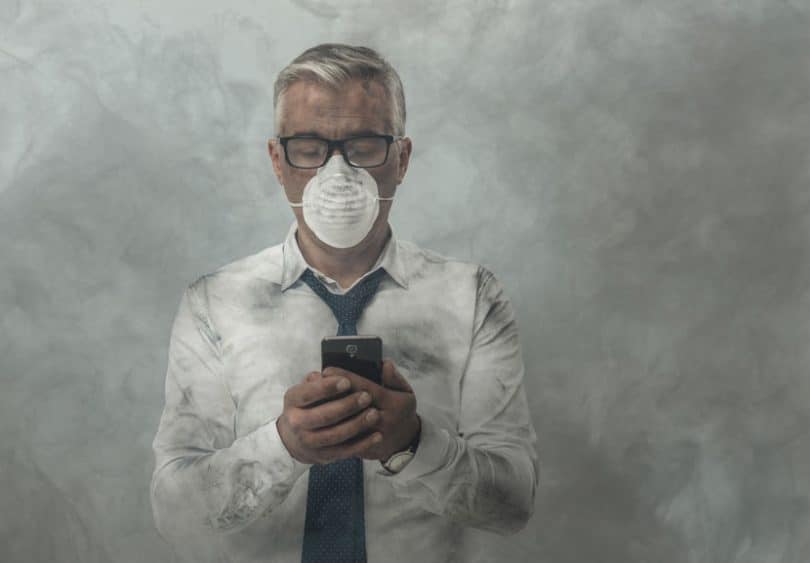Homem mexendo no celular com máscara no rosto e fumaça ao redor