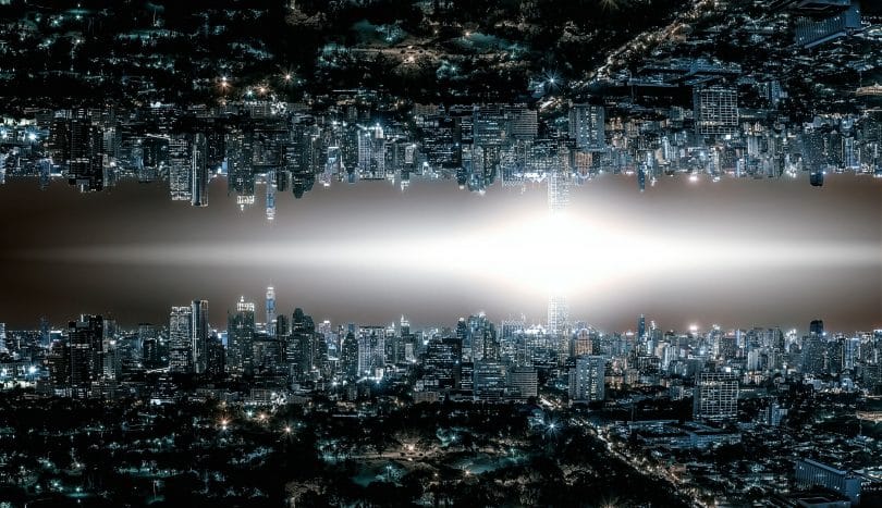 Horizonte de uma cidade grande com muitas luzes acesas em prédios e nas ruas. No centro da imagem existe uma luz em formato de losango. Na parte superior, a mesma imagem da parte de baixo mas de ponta-cabeça, como um reflexo.