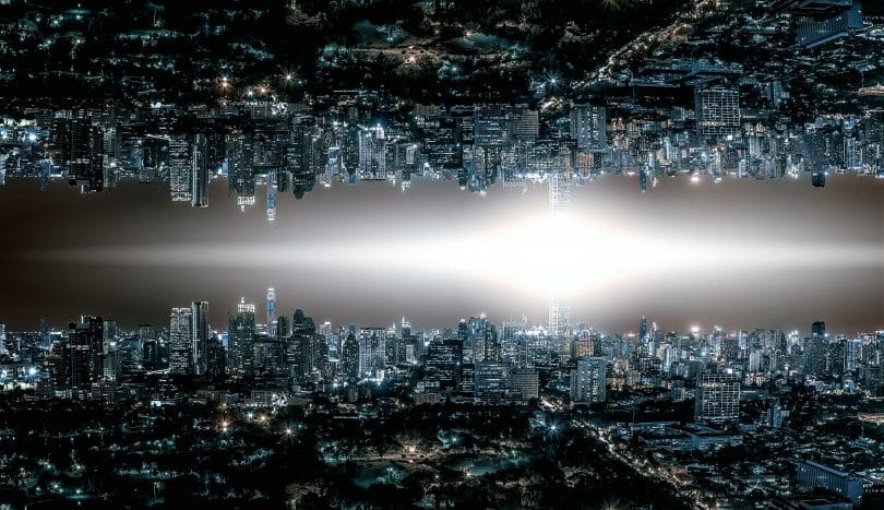 Horizonte de uma cidade grande com muitas luzes acesas em prédios e nas ruas. No centro da imagem existe uma luz em formato de losango. Na parte superior, a mesma imagem da parte de baixo mas de ponta-cabeça, como um reflexo.