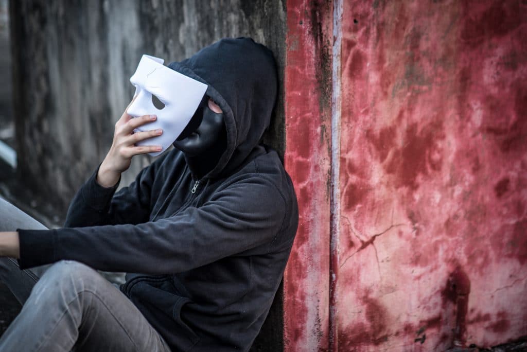 Homem vestindo um moletom preto e uma calça jeans cinza claro. Ele está sentado e encostado na parede. Ele usa em seu rosto uma máscara preta e em uma de suas mãos ele segura uma máscara branca sendo posta sobre a máscara preta.