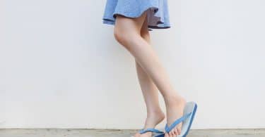Pernas de mulher vestindo saia e chinelos de dedo em frente a uma parede. Ela está de lado e uma de suas pernas está levemente flexionada, pois está apoiando na ponta do pé.