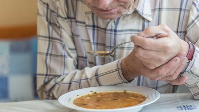 Imagem de um senhor de idade na frente de um prato de sopa levando uma colher para a boca com as duas mãos