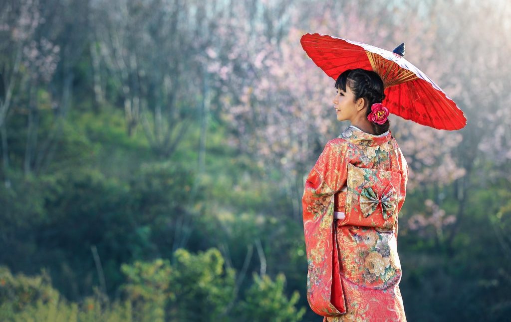 Imagem de uma linda japonesa usando uma sombrinha e um lindo kimono vermelho estampado com muitas flores. Ela está obserando uma linda floresta.