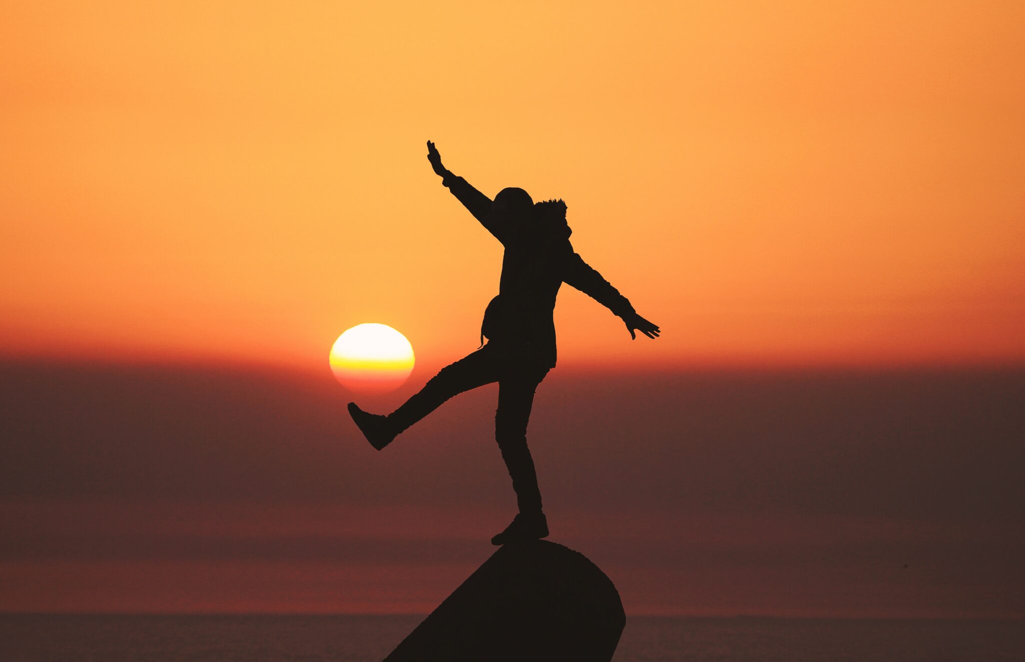 Silhueta de um homem em pé em um lugar alto, à frente de uma paisagem com pôr do sol. O homem tem seus braços abertos e um dos pés para a frente.