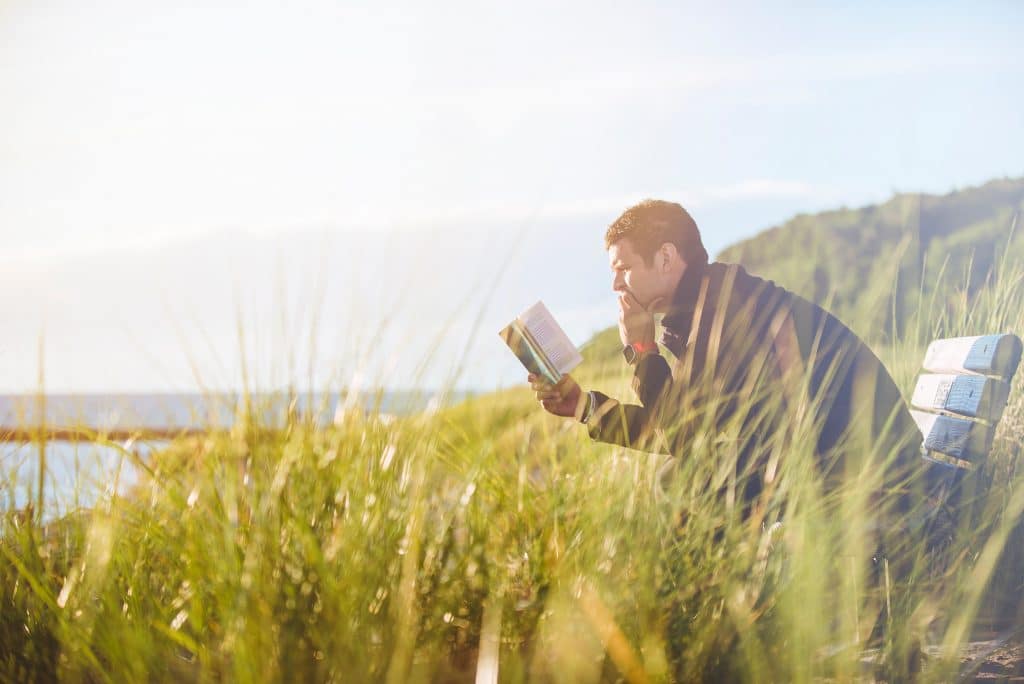 Homem sentado lendo um livro em uma paisagem natural, com um rio ao fundo.