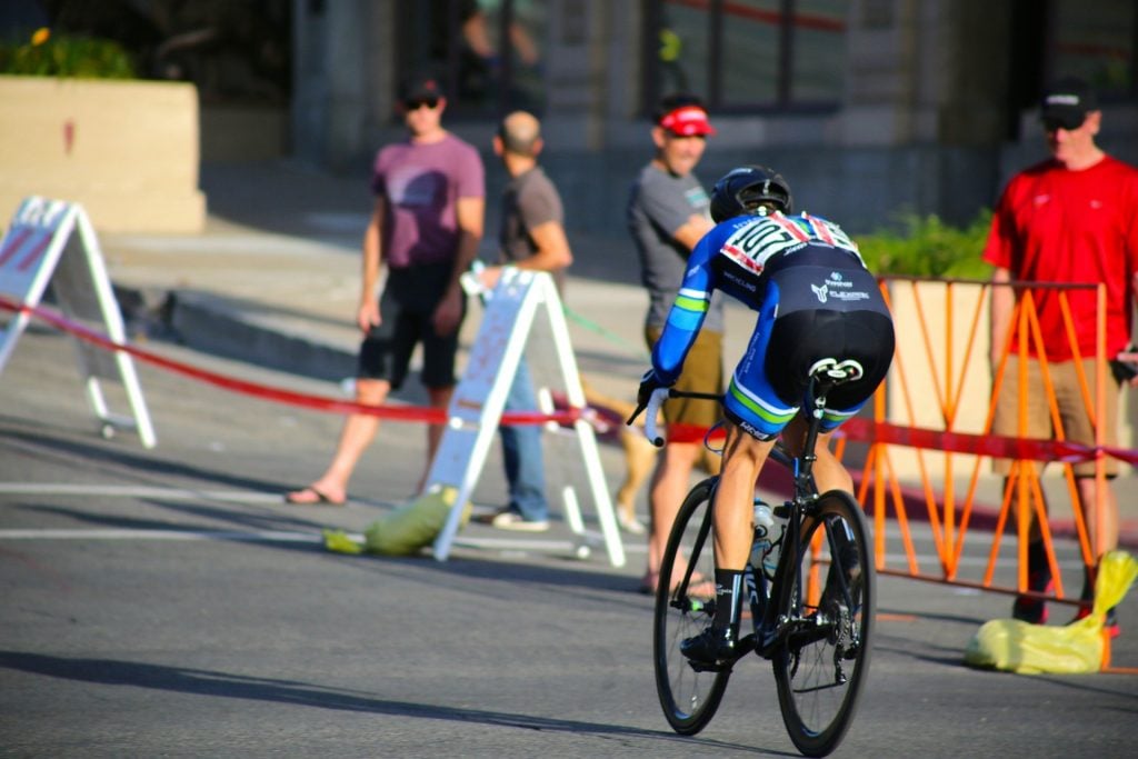 Homem praticando uma corrida de bicicleta. Ele usa roupas apropriadas para a prática desse esporte. Fora da pista de corrida, outros homems observam a corrida.
