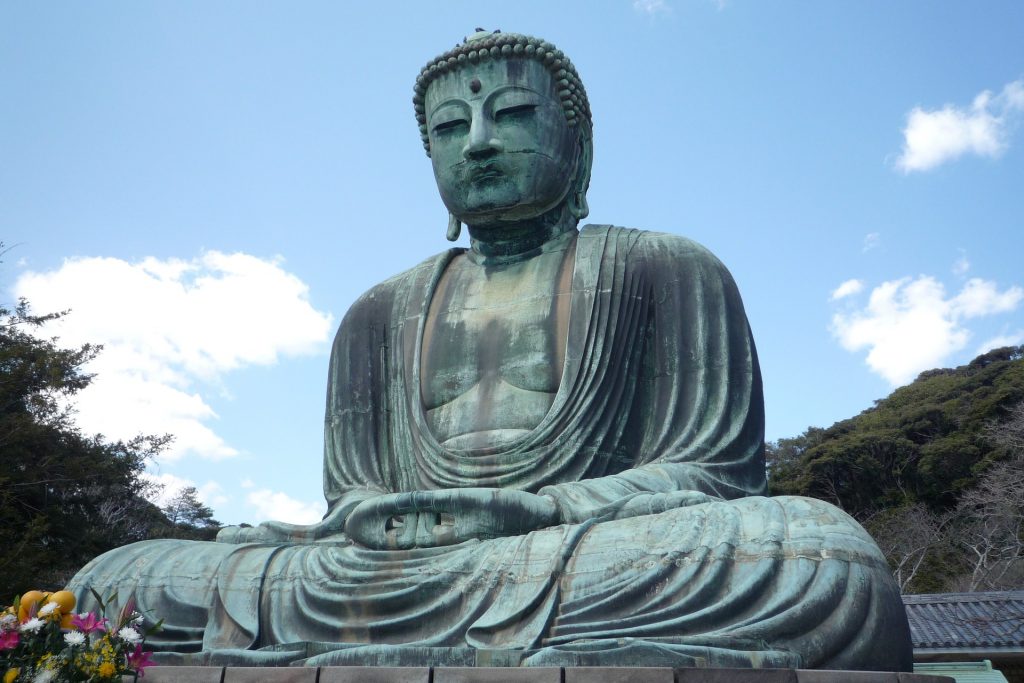 Linda imagem de uma estátua gigante de Buda em posição de meditação. Ao lado da estátua, vários arranjos de flores entregues como oferendas.
