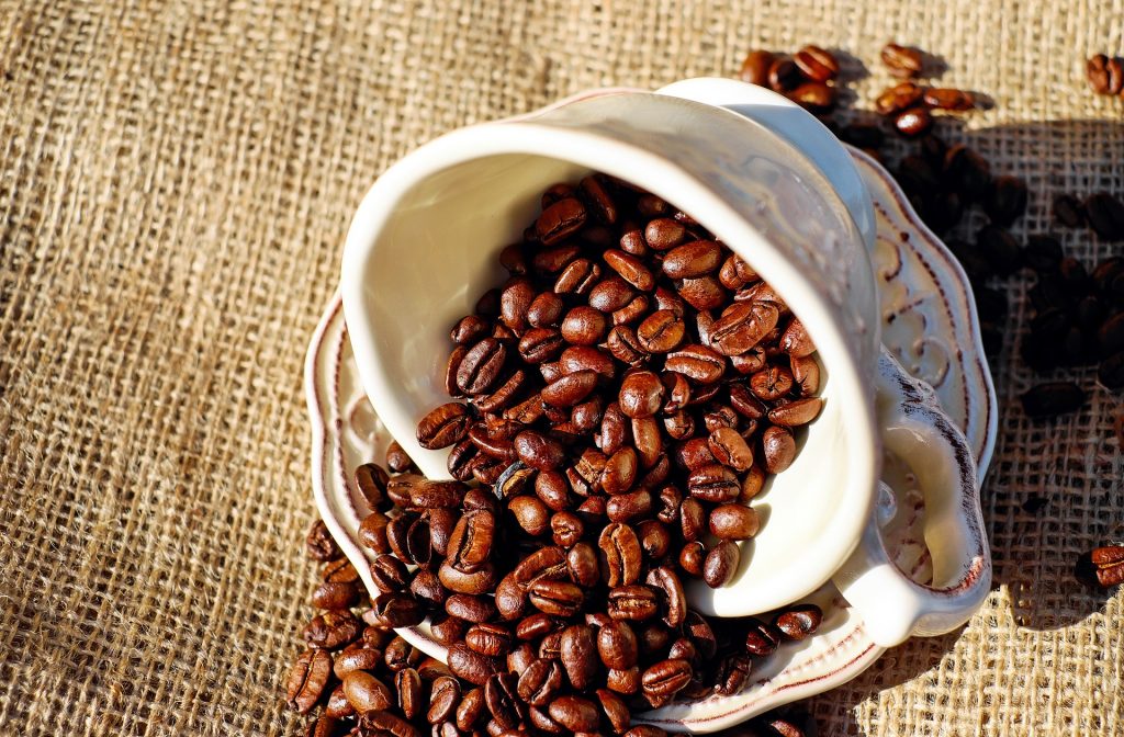 Imagem de um lindo conjunto de xícara e pires em porcelana. A xícara está cheia de grãos de café. O conjunto está sobre uma mesa forrada com um tecido de juta.
