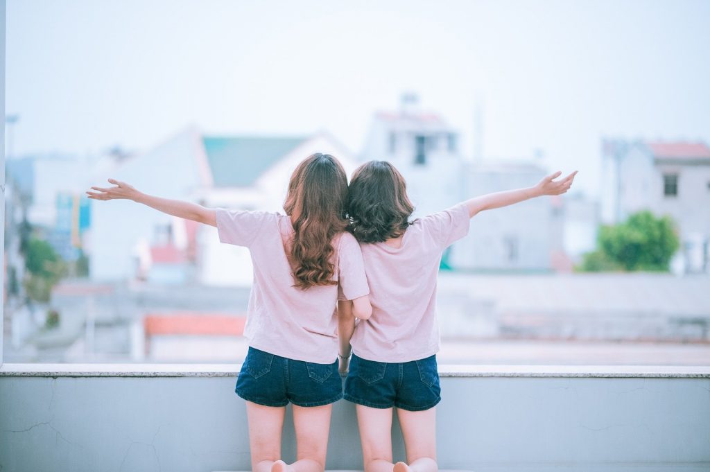 Imagem de um casal hetero. Elas estão de braços dados olhando para o infinito. Ambas estão vestidas iguais: shorts jeans escurto e camiseta branca.
