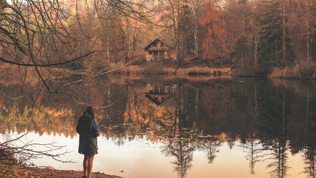 Mulher de costas, em pé, na frente de um lago cercado por árvores de outono, cujas folhas já estão quase todas caídas. Ao fundo, uma pequena casa é vista à beira do lago.