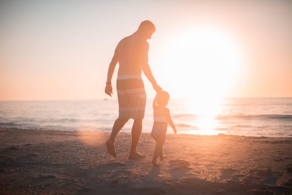 Homem adulto e criança pequena de mãos dadas, caminhando na praia em direção ao mar.