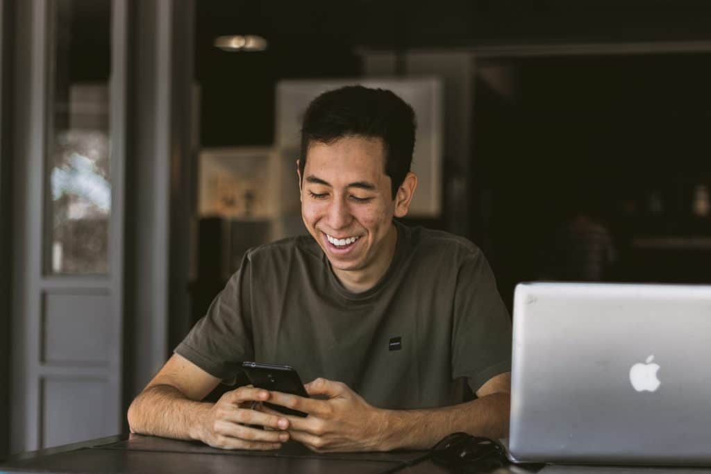 Homem sentado, sorrindo, enquanto usa seu smartphone.