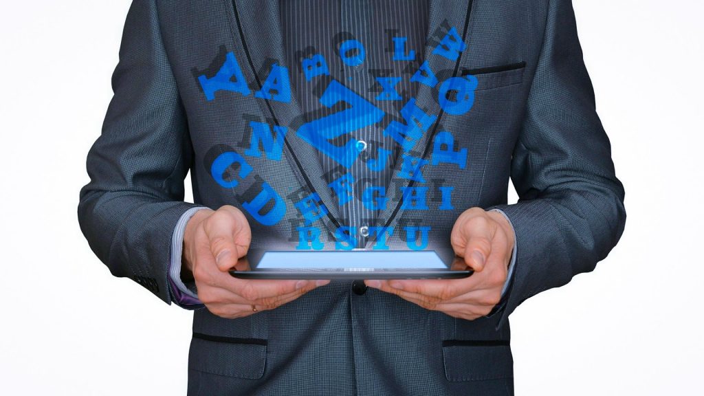 Homem vestindo um terno azul. Ele segura em suas mãos um aparelho eletrônico. Ao fundo imagens de letras do alfabeto em cor azul claro compõem a imagem.