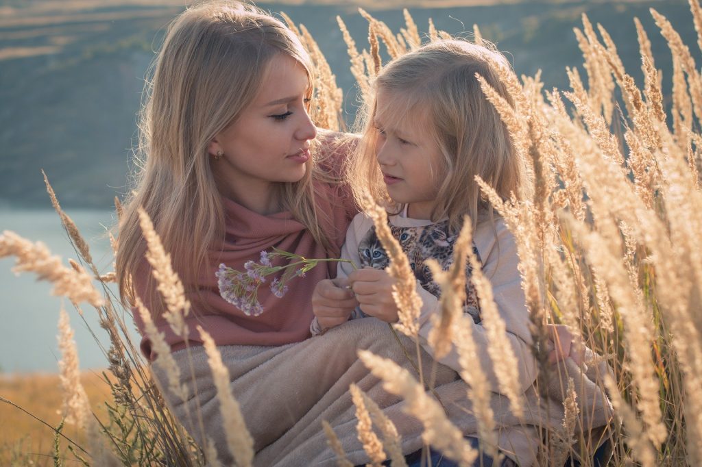 Imagem de mãe e filha em um lindo dia ensolarado. A criança está sentada no colo da mãe que está sentada em um campo com uma plantação de folhagens douradas.