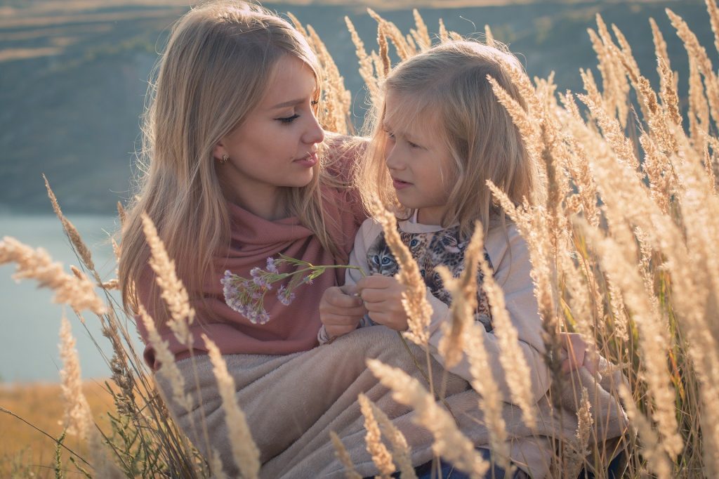 Imagem de mãe e filha em um lindo dia ensolarado. A criança está sentada no colo da mãe que está sentada em um campo com uma plantação de folhagens douradas.
