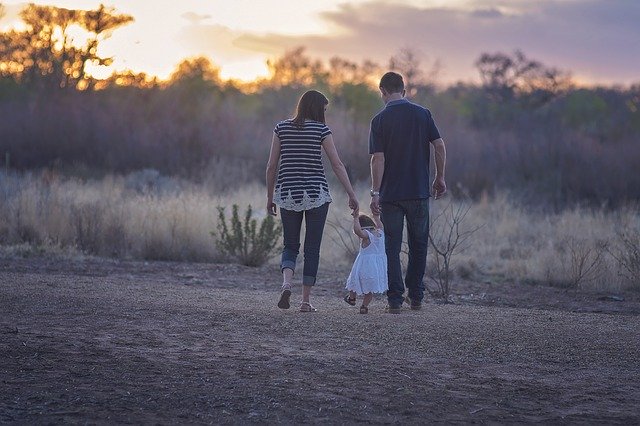 Mãe e pai de mãos dadas com filhinha aprendendo a andar em campo ao entardecer