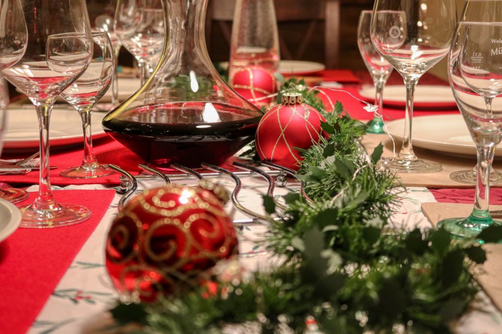 Imagem de uma linda mesa de Natal. Ela está decorada com tolhas branca e vermelha, enfeites natalinos e sobre a mesa um cantil de vidro com vinho e várias taças.