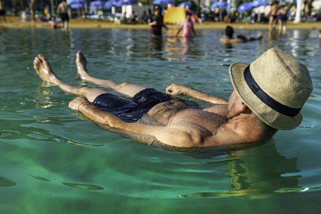 Homem flutuando em um lago. Ele usa uma bermuda azul e  um chapéu de palha com uma fita preta. No lago também se encontram outras pessoas.
