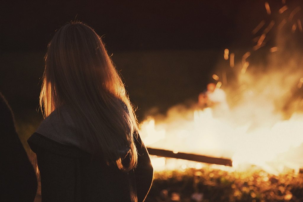Imagem de uma mulher loura vestindo uma blusa de frio com touca. Ela está olhando para o fogo.