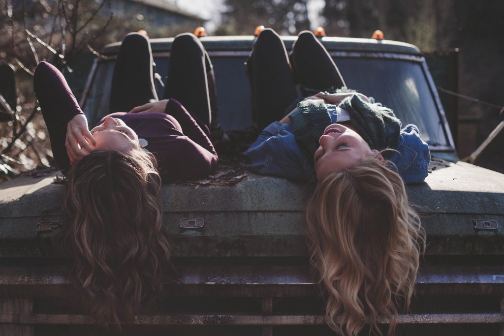 Imagem de duas garotas de cabelos longos, deitadas sobre a caçamba de uma caminhonete antiga. Elas estão felizes.