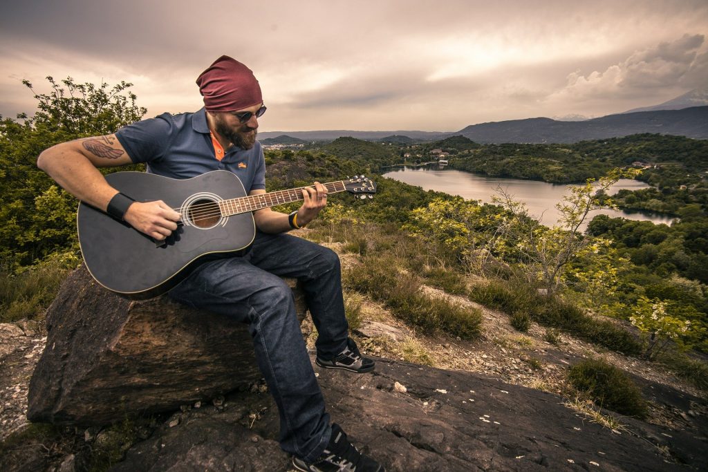 Imagem de um homem em um vale. Ele está sentado sobre uma pedra. Veste roupas jeans, azul escuro e usa um óculos escuro. Sobre a cabeça uma bandana vermelha. Ele está tocando um violão.

