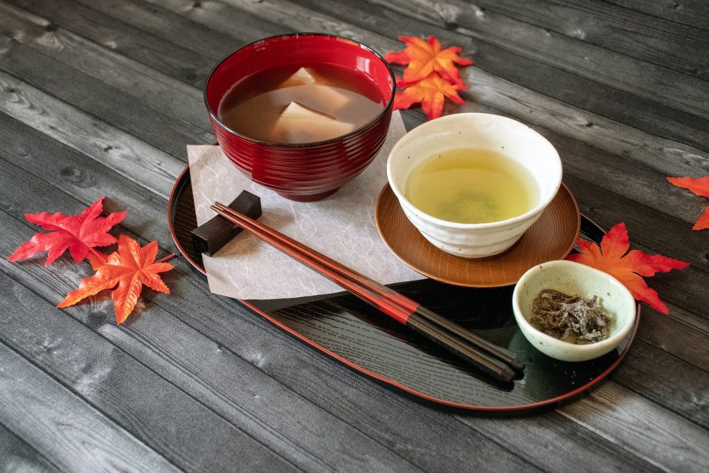 Mesa posta com vários itens para a reallização da cerimônia do chá japonesa. Os itens estão sobre uma mesa de madeira cinza decorado com folhas de outono na cor vermelha.
