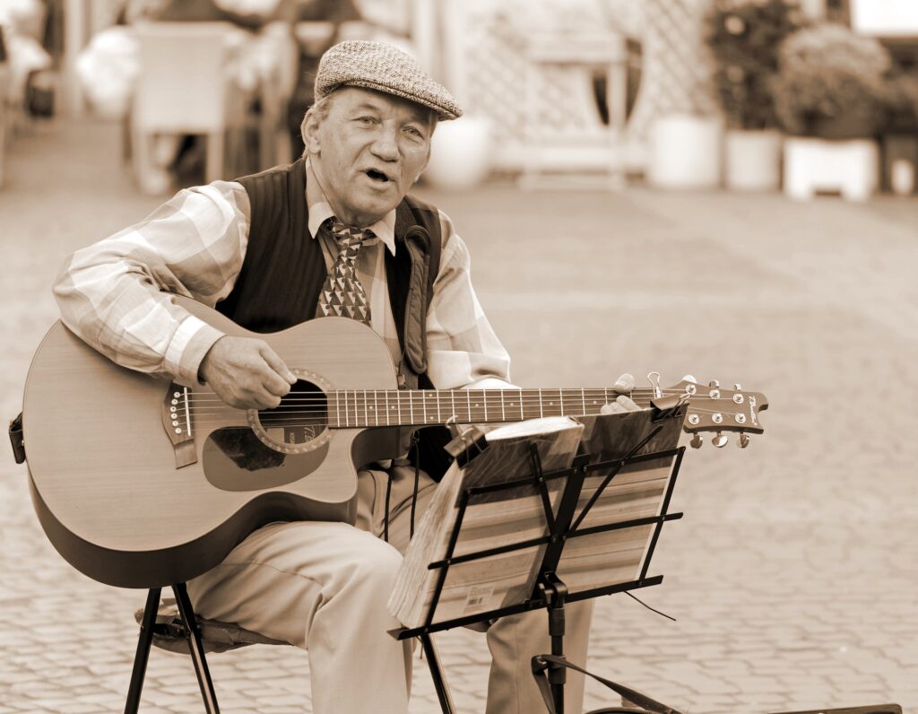 Imagem em preto e branco e um senhor usando boina e colete, sentando em uma praça. Ele está cantando e tocando um violão.
