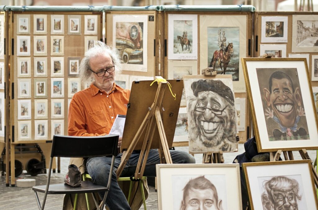 Homem idoso pintando um quadro em uma feira, com diversas exposições de desenhos e pinturas.