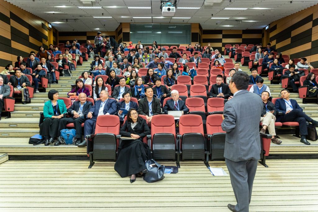 Plateia em auditório assistindo palestra, de homem que fala de costas para a câmera.