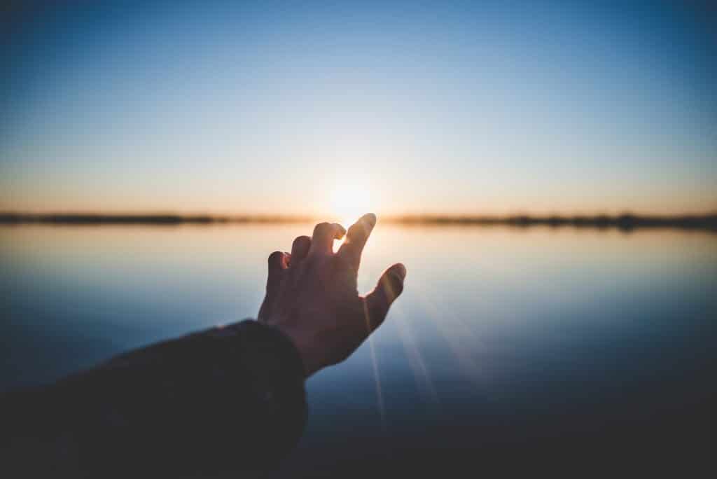 Mão estendida em frente a um rio, em direção ao sol, que se põe.