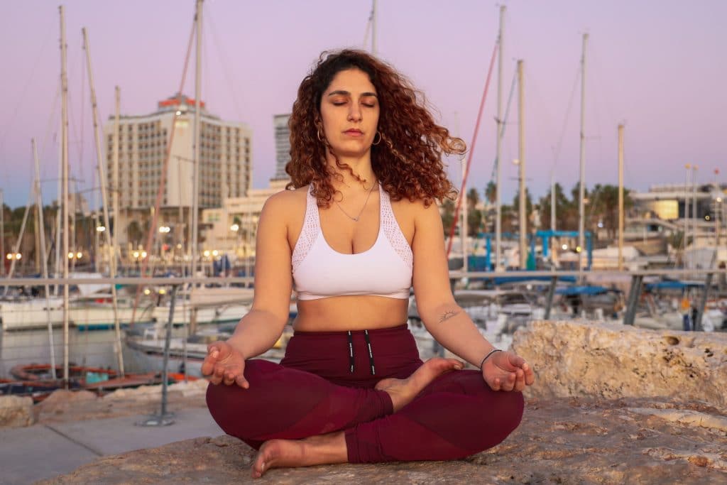 Mulher sentada em uma pedra em posição de meditação e olhos fechados.  Atrás há um porto com diversos barcos.