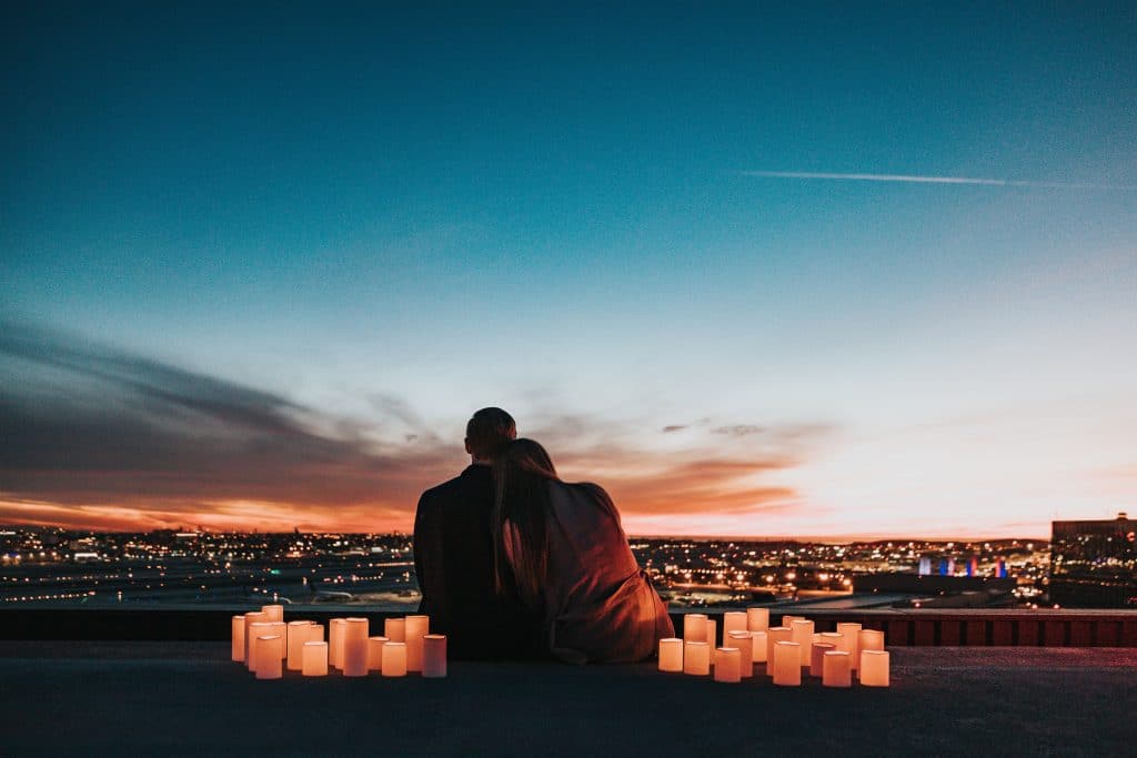 Casal sentado no topo de um prédio, rodeado por velas acesas, ao pôr do sol.