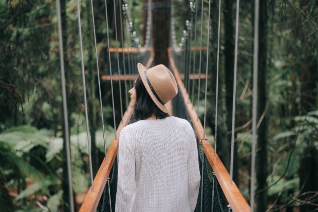 Imagem de uma mulher atravessando um ponte no meio de uma floresta. Ela está usando roupas claras e um chapéu clássico na cor bege.
