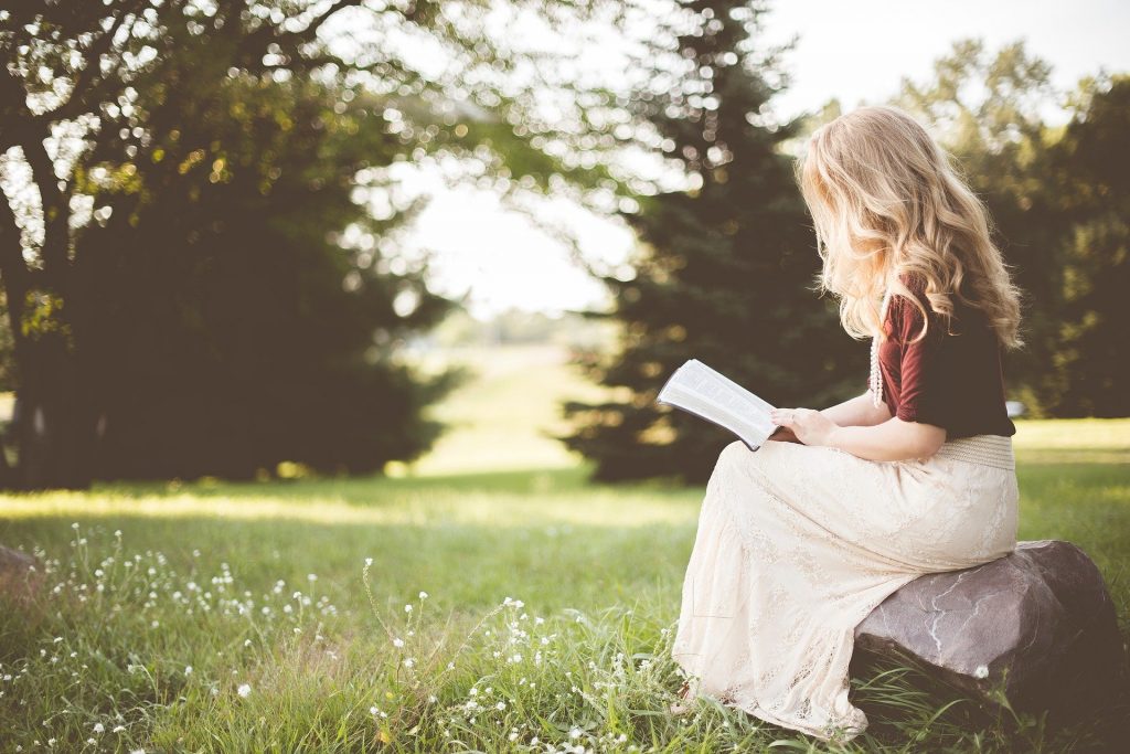 Imagem de um lindo campo verde. Nele encontra-se uma linda mulher loura de cabelos longos. Ela está sentada sobre uma pedra e fazendo a leitura de um livro.
