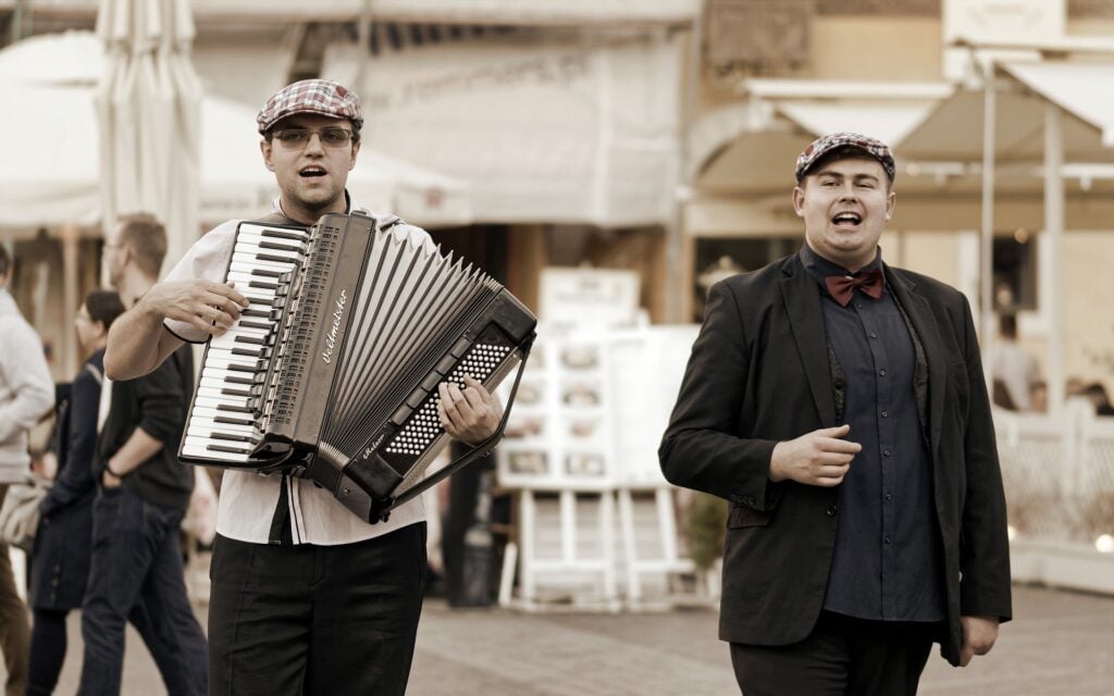 Dois homens jovens usando boina e roupa social. Eles estão em uma praça aberta. Ambos estão cantando e um deles tocando sanfona.
