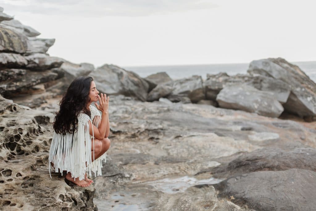 Mulher agachada com olhos fechados em pedras com mar ao fundo